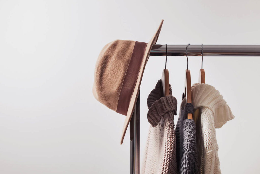 Three fall sweaters hanging on a rolling rack with a brown hat hanging on the side of the rack